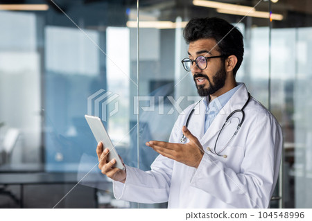Successful professional Indian doctor working inside modern clinic, man consulting patients remotely, using tablet computer for video call, and long distance communication. 104548996