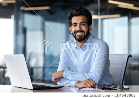 Portrait of happy and successful businessman, indian man smiling and looking at camera, satisfied with achievement results man working inside office building using laptop at work. 104549002