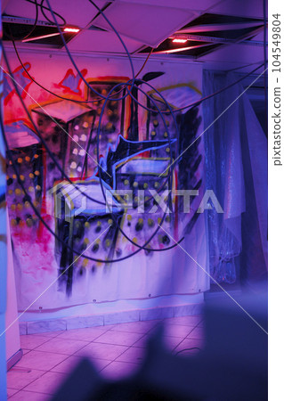 Deserted urban space with purple and pink neon lights, showing dust on walls, abandoned warehouse with crumbling structure covered in messy graffiti paint. Place with fluorescent lights. 104549804