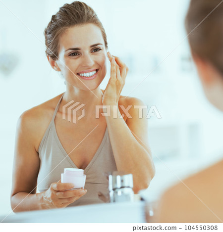 Happy young woman applying cream in bathroom Happy young woman applying cream in bathroom 104550308