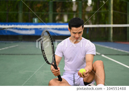 Handsome Asian male tennis player in white sport clothes holding ball racket sitting on court 104551488