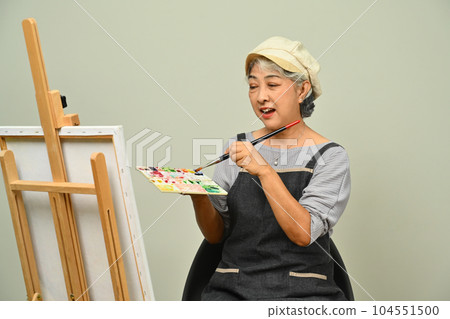 Portrait of gray haired senior woman wearing apron holding palette painting picture over gray background 104551500