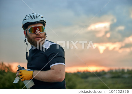 Close-up portrait of young bearded man, cyclist in helmet and special glasses standing over sunset sky background Close-up portrait of young bearded man, cyclist in helmet and special glasses standing over sunset sky background 104552856
