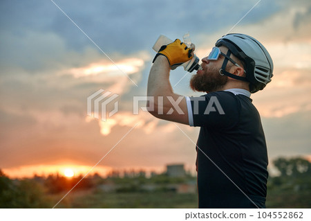 Young sportive man, cyclist in helmet and comfortable clothes standing by the road with bike and drinking water. Sunset 104552862