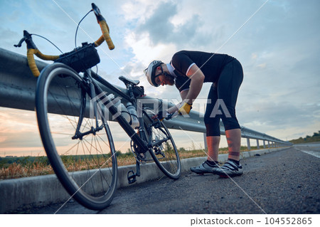 Young bearded man, cyclist in sportswear and helmet, pumping up bicycle wheel on road. Riding in chill evening Young bearded man, cyclist in sportswear and helmet, pumping up bicycle wheel on road. Riding in chill evening 104552865