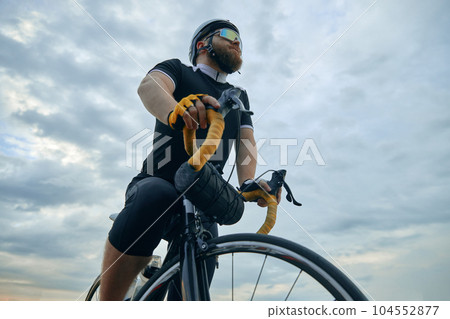 Bottom view image of sportive bearded man in sportswear, glasses and helmet standing with bicycle over sky background Bottom view image of sportive bearded man in sportswear, glasses and helmet standing with bicycle over sky background 104552877