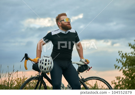 Handsome, young, bearded man in sport glasses and sportswear, cyclist sitting in bike over field background Handsome, young, bearded man in sport glasses and sportswear, cyclist sitting in bike over field background 104552879