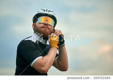 Young bearded man, bike rider in gradient sport glasses, putting on helmet over cloudy sky background Young bearded man, bike rider in gradient sport glasses, putting on helmet over cloudy sky background 104552884