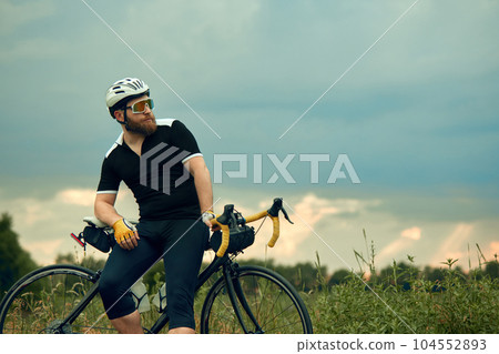 Bearded man in sportswear, glasses and helmet, sitting on bike by the road on field in chill summer evening Bearded man in sportswear, glasses and helmet, sitting on bike by the road on field in chill summer evening 104552893
