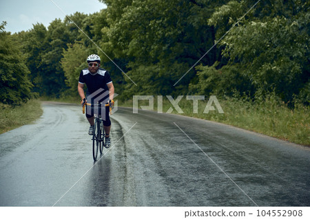 Sportive, young, bearded man, cyclist in helmet, glasses and uniform riding bike on wet road in cloudy chill evening Sportive, young, bearded man, cyclist in helmet, glasses and uniform riding bike on wet road in cloudy chill evening 104552908