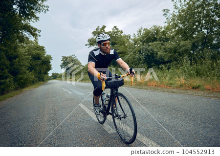 Young bearded man, cyclist in helmet, glasses and uniform riding bike on wet road in cloudy chill evening 104552913