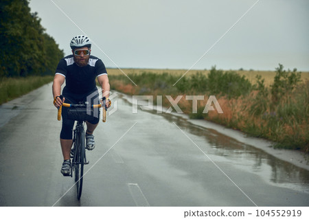 Bearded young man, cyclist in helmet, glasses and uniform riding bike on wet road in cloudy chill evening 104552919