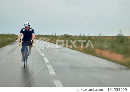 Fast ride. Young bearded man, cyclist in helmet, glasses and uniform riding bike on wet road in cloudy chill evening Fast ride. Young bearded man, cyclist in helmet, glasses and uniform riding bike on wet road in cloudy chill evening 104552929