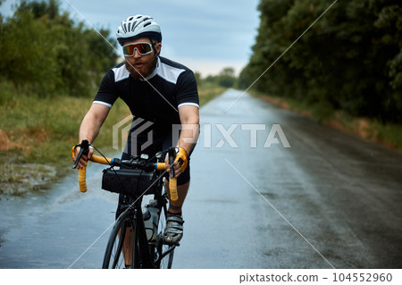 Concentrated, young, bearded man, cyclist in uniform, helmet and glasses riding bike on road in chill evening. 104552960