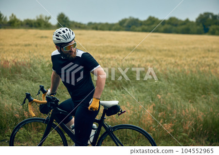 Bearded young man, athlete cyclist in uniform, helmet and glasses sitting on bicycle by the road. Field background Bearded young man, athlete cyclist in uniform, helmet and glasses sitting on bicycle by the road. Field background 104552965
