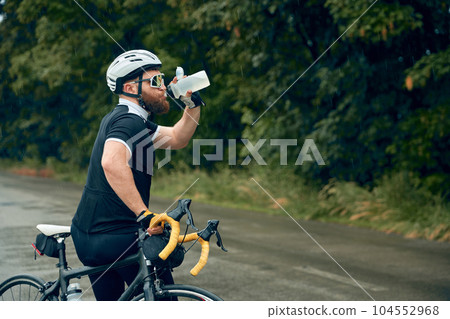 Bearded young man, cyclist in uniform, helmet and glasses standing by the road and drinking water. Refreshment and rest 104552968