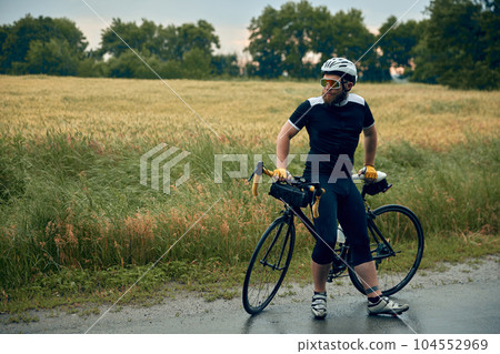 Young bearded man, cyclist in uniform, helmet and glasses sitting on bicycle on road around field background 104552969