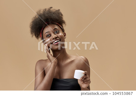 Portrait of young, positive, beautiful, african woman applying face moisturizing cream against studio background Portrait of young, positive, beautiful, african woman applying face moisturizing cream against studio background 104553867