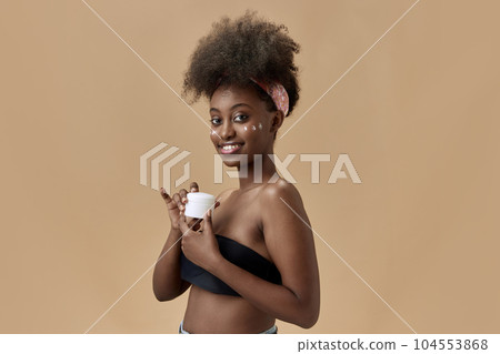 Organic cosmetics. Portrait of young, beautiful, african woman applying face moisturizing cream, spf against studio background Organic cosmetics. Portrait of young, beautiful, african woman applying face moisturizing cream, spf against studio background 104553868