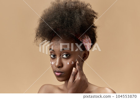 Close-up portrait of young, beautiful, african woman applying face moisturizing cream against studio background 104553870