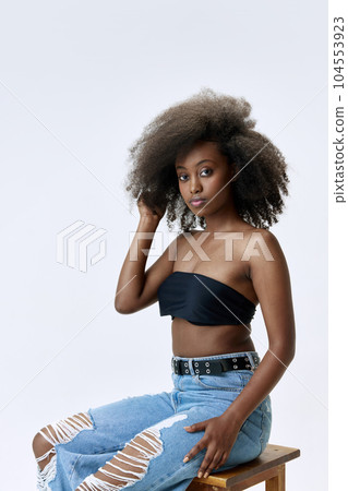 Portrait of beautiful, attractive, young african girl with well-kept skin, curly hair, sitting in jeans and top against white studio background 104553923