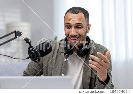 Close-up photo. Young hispanic man sitting at home wearing headphones, talking into a microphone on a video call from a laptop. He looks smilingly at the monitor, communicates. Close-up photo. Young hispanic man sitting at home wearing headphones, talking into a microphone on a video call from a laptop. He looks smilingly at the monitor, communicates. 104554024