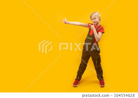 Blond boy wears jumpsuit and red T-shirt stands with his legs spread wide and pointing his index fingers on side. Copy space. Blond boy wears jumpsuit and red T-shirt stands with his legs spread wide and pointing his index fingers on side. Copy space. 104554828