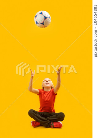 Boy sits cross-legged and tosses soccer ball up in isolation against yellow background. Vertical frame. 104554893