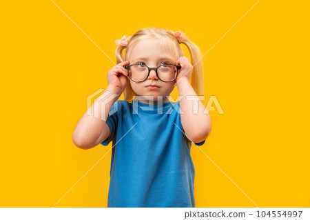 Elementary schoolgirl with two ponytails wears blue T-shirt and glasses looks up. Studio portrait of child, yellow background Elementary schoolgirl with two ponytails wears blue T-shirt and glasses looks up. Studio portrait of child, yellow background 104554997
