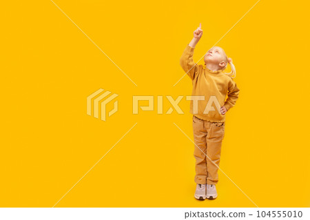 Child raised hand and points her finger up. Studio portrait of girl in yellow suit on yellow background. Space for text 104555010