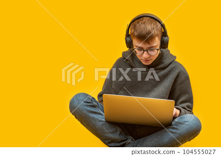 Teenage boy in jeans hoodie glasses and headphones sits cross-legged holding laptop. Online learning. Programming for children. Copy space. Teenage boy in jeans hoodie glasses and headphones sits cross-legged holding laptop. Online learning. Programming for children. Copy space. 104555027