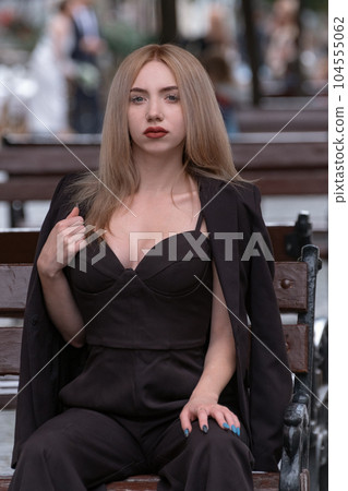 Portrait of serious young attractive blonde woman in black suit with deep neckline on bench. Vertical frame 104555062