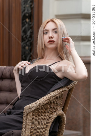 Young blonde beauty wears black top bustier with deep neckline sits in wicker chair and looks into the camera. 104555063