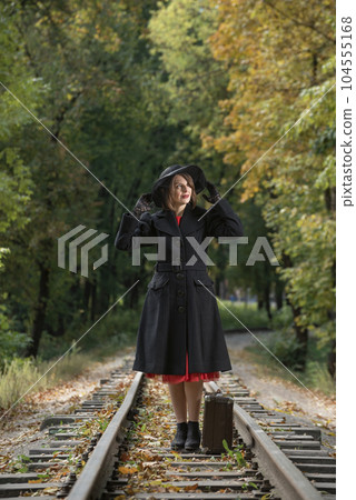 Woman in retro clothes black coat and broad-brim hat stands with old valise on rails. Woman dressed in 40s style. 104555168