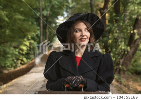 Portrait woman in black hat with brims, classic jaket and gloves rests hands on old suitcase. Forest background Portrait woman in black hat with brims, classic jaket and gloves rests hands on old suitcase. Forest background 104555169