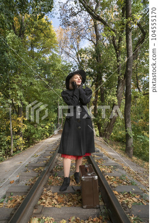 Lady in stylish black coat and broad-brim hat stands dreamily near vintage suitcase on rails in autumn wood. Vertical frame 104555170