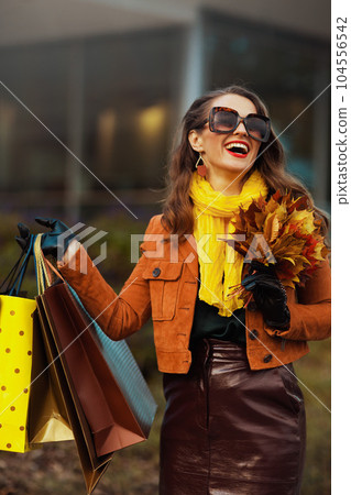 smiling trendy woman in brown trench coat with shopping bags 104556542