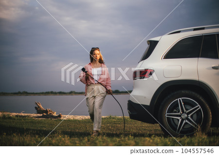 Young woman holding power supply cable from her electric car, prepared for charging it sustainable and economic transportation concept. Young woman holding power supply cable from her electric car, prepared for charging it sustainable and economic transportation concept. 104557546