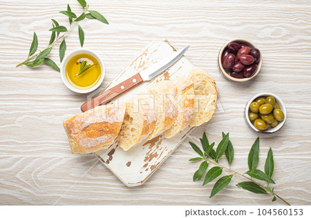 Sliced fresh ciabatta on cutting kitchen board, green and brown olives, olive oil with rosemary, olive tree branches on white wooden rustic background top view 104560153