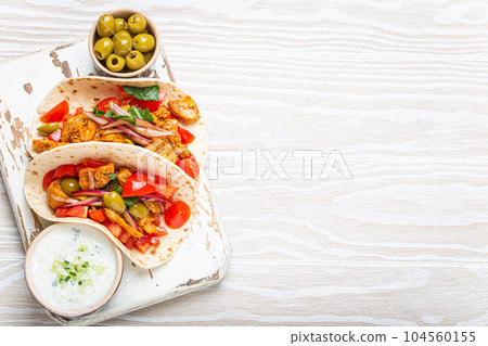 Traditional Greek Dish Gyros: Pita bread Wraps with vegetables, meat, herbs, olives on rustic wooden cutting board with Tzatziki sauce, olive oil top view on white wooden background, space for text. 104560155