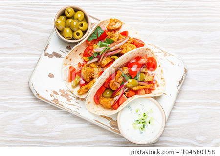 Traditional Greek Dish Gyros: Pita bread Wraps with vegetables, meat, herbs, olives on rustic wooden cutting board with Tzatziki sauce, olive oil top view on white wooden summer background. 104560158