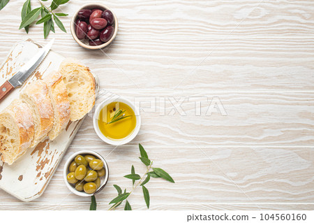 Sliced fresh ciabatta on cutting kitchen board, green and brown olives, olive oil with rosemary, olive tree branches on white wooden rustic background top view 104560160