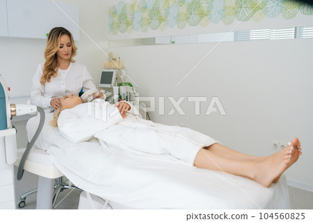 Wide shot of professional female cosmetician performing photo rejuvenation cosmetology procedure for young woman in modern cosmetology clinic. Concept of anti-age rejuvenation skincare. Wide shot of professional female cosmetician performing photo rejuvenation cosmetology procedure for young woman in modern cosmetology clinic. Concept of anti-age rejuvenation skincare. 104560825