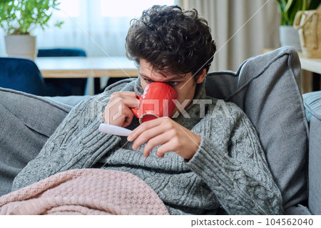 Young guy checking temperature, lying on couch under blanket 104562040