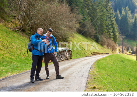 Two hikers are in the mountains in the spring nature outdoor. Hiking with backpacks. Two friends travel together in picturesque places. The man shows the way. Two men on a hiking trip in the mountains Two hikers are in the mountains in the spring nature outdoor. Hiking with backpacks. Two friends travel together in picturesque places. The man shows the way. Two men on a hiking trip in the mountains 104562358