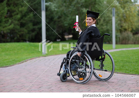 Happy caucasian woman in a wheelchair turns around and holds her diploma outdoors. Happy caucasian woman in a wheelchair turns around and holds her diploma outdoors. 104566007