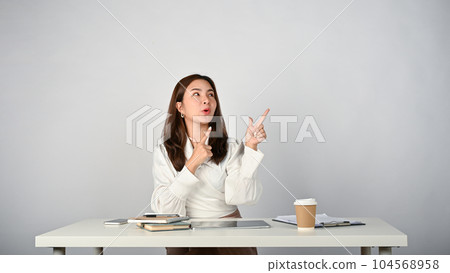 A cheerful young Asian businesswoman  looking up and pointing her fingers up at empty space. 104568958