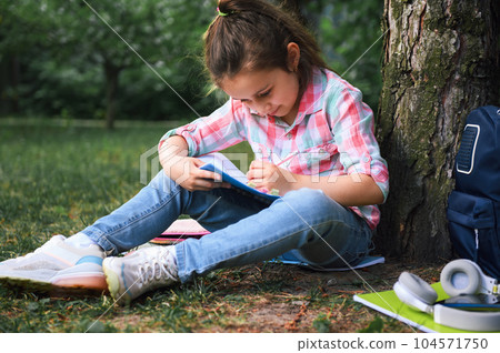 Adorable child girl, primary school student in stylish checkered shirt and blue denim, doing homework in the city park. 104571750