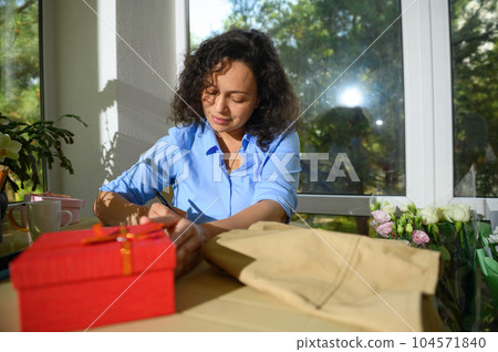 Beautiful curly haired Latin American woman taking notes in a floral workshop. Beige apron and red gift box on the table Beautiful curly haired Latin American woman taking notes in a floral workshop. Beige apron and red gift box on the table 104571840