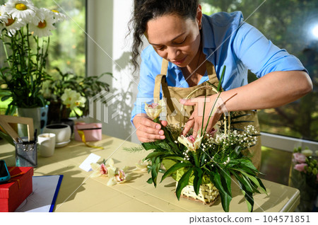 Female florist arranging a flower bouquet, inserting orchid flower into a soaked foam in a wicker basket in floral shop Female florist arranging a flower bouquet, inserting orchid flower into a soaked foam in a wicker basket in floral shop 104571851